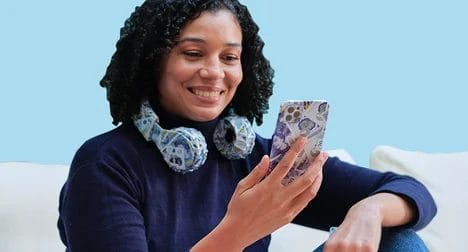 Medium-dark skinned woman sitting on a sofa smiling at her smart phone and headphones which are both wrapped in £5, £10 and £20 notes.