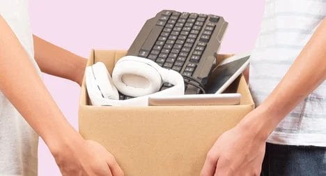 Two light skinned people holding a cardboard box full of old electrical items against a pink background.