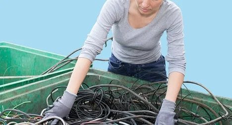 Light skinned young woman sorts cables at waste centre, wearing gloves against a bright blue background.