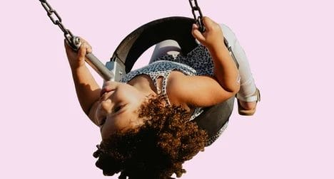 A medium skinned child on a tyre swing, mid-swing upside down, against a pink background.