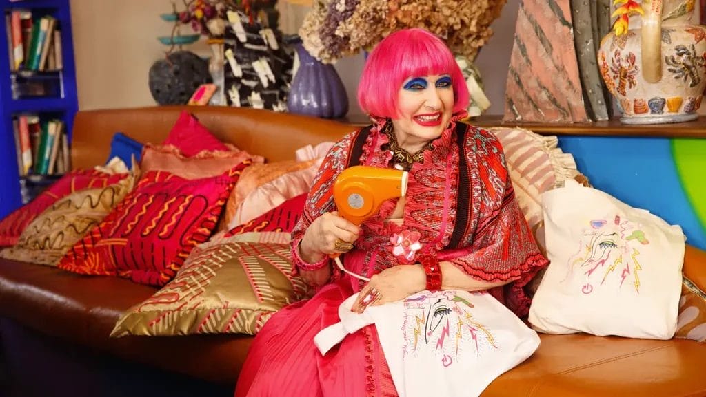 Light skinned fashion designer Zandra Rhodes wearing pink with pink hair holding electrical tote bag she designed and a hairdryer, sitting on a sofa with bright cushions