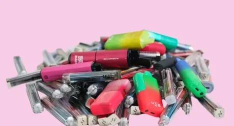 many multi-coloured and two-tone colour vapes scattered on a pink table top