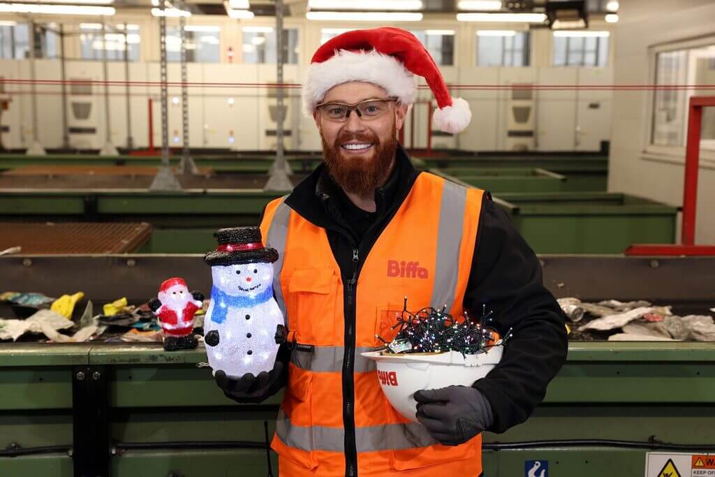 A recycling worker wearing a high-visibility orange vest and Santa hat stands inside a recycling facility, holding a light-up snowman decoration and a hard hat filled with tangled Christmas lights, with a conveyor belt behind them.