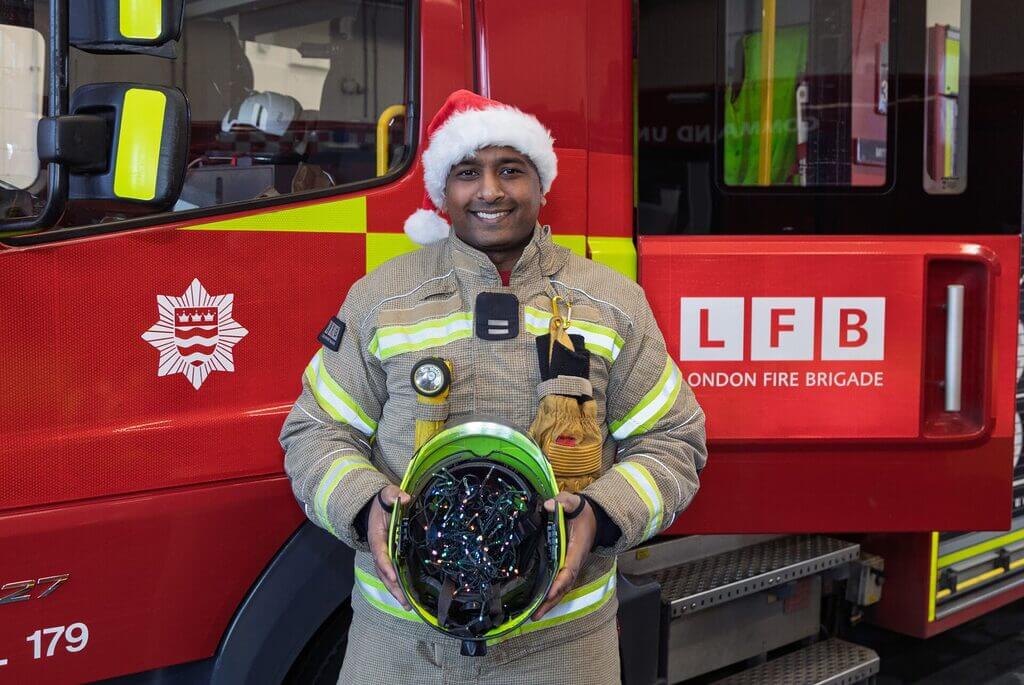 A firefighter wearing full protective gear and a Santa hat stands in front of a London Fire Brigade fire engine, holding a helmet filled with tangled Christmas lights.