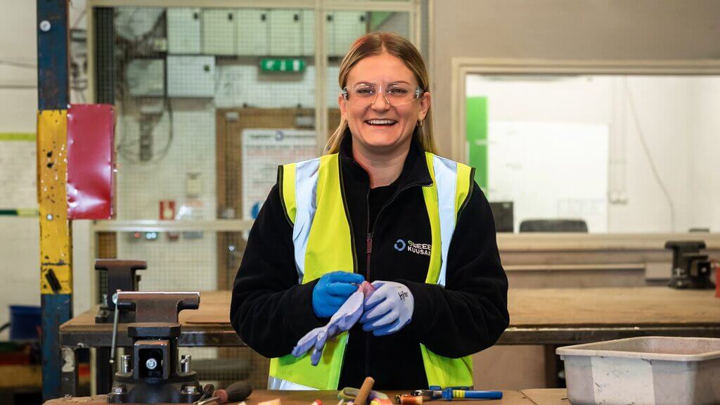 Alt=""A recycling worker wearing a high-visibility yellow vest, safety glasses, and gloves smiles while standing at a workbench in a workshop, with tools and equipment visible around them.