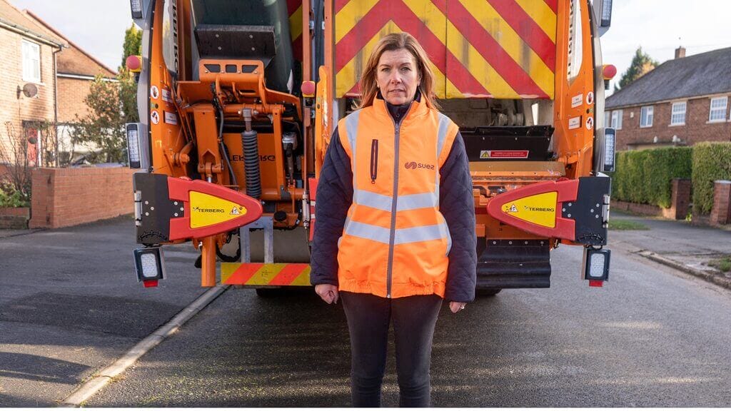 A waste collection worker wearing a high-visibility orange vest stands on a residential street in front of a refuse truck, facing the camera with houses visible in the background.
