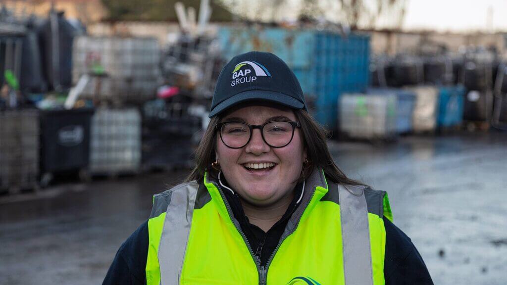 Alt=""A smiling recycling worker wearing a high-visibility vest and cap stands at an outdoor recycling facility, with large containers and equipment behind them.