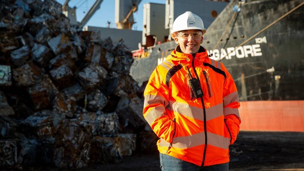 A worker wearing a high-visibility orange jacket and white hard hat stands smiling in front of stacked metal scrap at a dock, with a large cargo ship in the background.