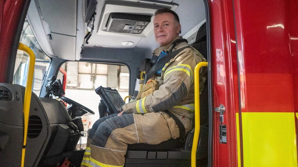 A firefighter wearing full protective gear sits in the cab of a fire engine, facing the camera
