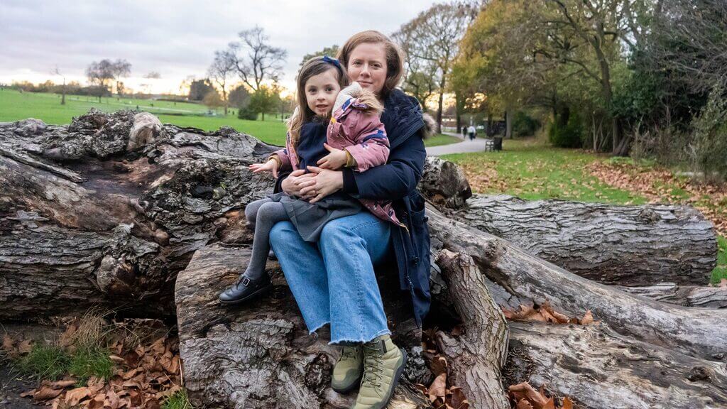 An adult sits on a fallen tree trunk in a park holding a young child, with autumn leaves on the ground and open green space in the background.