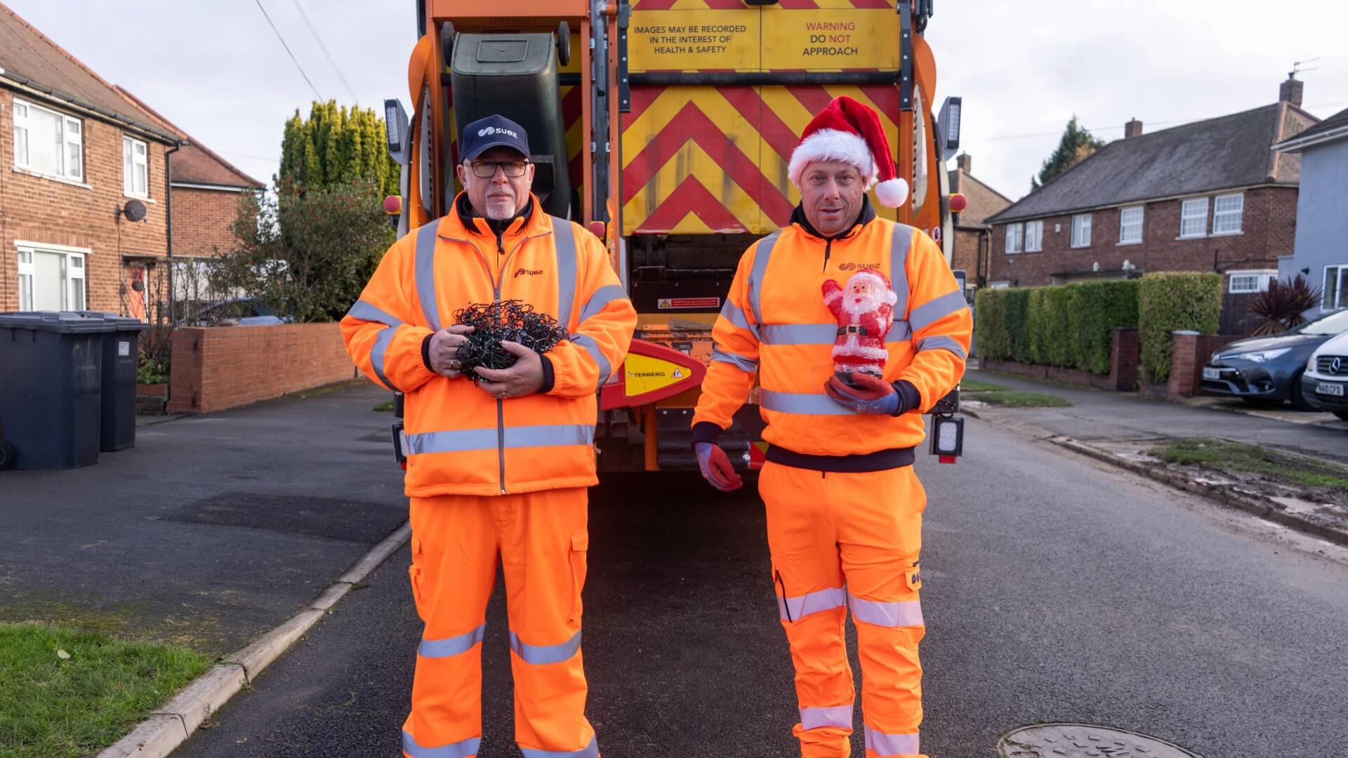 Two waste collection workers in high-visibility orange clothing stand on a residential street in front of a refuse truck, holding a bundle of tangled string lights and a festive ornament.